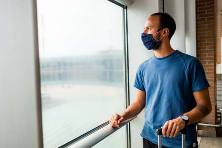 young man wearing protective face mask, traveling in the time of covid19 pandemic inside an airport, looking out of window with worried and upset expression. corona virus and travel industryの写真素材