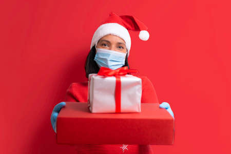 covid christmas concept. young smiling woman holding boxes of gifts, face mask, santa claus outfit smiling on red plain background. christmas during covid19 pandemic.の写真素材