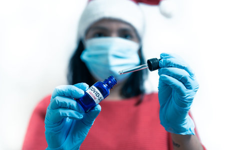 covid-19 vaccine christmas concept. female doctor with face mask in santa claus outfit holding covid...