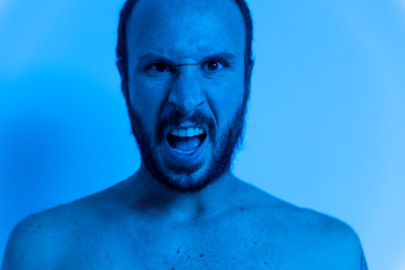 naked angry man screaming. waist level close up of furious young male with rage on blue background with beard. concept of stress and mental health in young adult. abusive toxic masculinityの写真素材