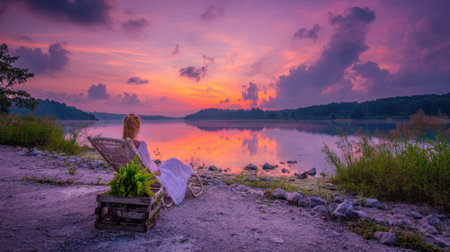 Woman sits peacefully on a sun lounger at the edge of a calm lake, as the sun rises, casting a warm light over the serene landscape.  The tranquil scene is filled with the soft colors of the sunrise reflecting on the water's surface.の素材
