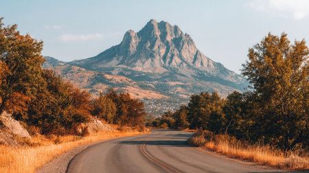 Winding asphalt road meanders through a valley lined with autumnal foliage, leading to a majestic mountain peak.  The colors of the trees are vibrant, blending with the light tan and grayish tones of the mountain's rock face.の素材