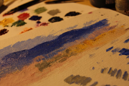 Close-up view of a painting palette with various colors and brushstrokes of paint applied to a white surface.  The palette shows a range of colors including blue, yellow, brown, pink, and gray, with discernible brushstrokes blending these hues. The overall impression is a work in progress, suggesting a stage of creative exploration.の素材