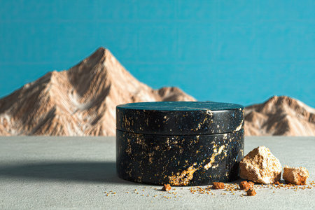 Dark, round container with golden speckles sits on a light-gray surface.  The container is positioned against a backdrop of beige mountains against a vibrant blue sky, and small rocks are scattered around the base of the container.の素材