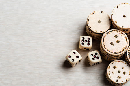 High-angle close-up view of several light-colored wooden dice and circular  chips stacked on a light-colored wooden surface. The dice and chips show various markings and numbers.の素材