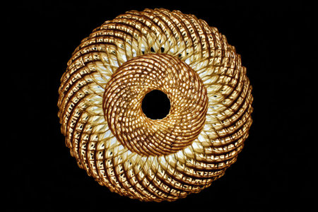 Circular, golden design composed of intricate, interwoven, spiral patterns resembling woven wicker or cane. The light from within illuminates the design, creating a captivating visual effect. The design is perfect for adding a touch of elegance to a room.の素材
