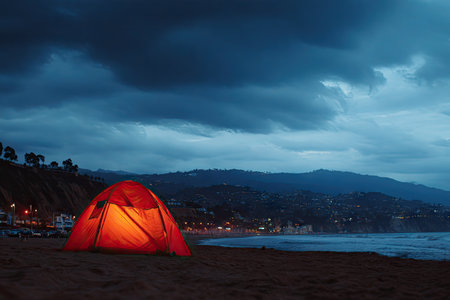 Red camping tent is lit up on a beach at twilight. The ocean waves meet the shore. Dark clouds and mountains are visible in the background. The scene has a serene and peaceful quality.の素材