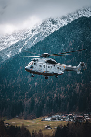 Helicopter is in flight over a valley and small village, with snow-capped mountains in the background. Lush green grass and pine trees are visible in the valley, creating a tranquil scene.の素材