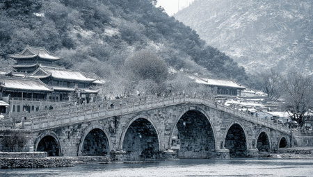Stone arch bridge spans a frozen river in a snowy, mountainous landscape.  Traditional Asian-style buildings are visible along the riverbanks. The image has a muted, monochromatic tone.の素材