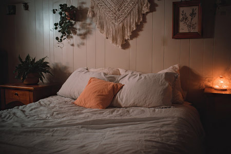 Bedroom scene featuring a bed with a soft, light-gray linen spread, comfortable pillows, an orange accent pillow, a wooden nightstand, and a plant.  Warm lighting creates a tranquil atmosphere.  The walls are a muted beige with simple wooden paneling, and there is a hanging woven wall decor and a framed piece of art on the wall.の素材