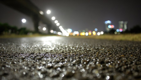 Close-up view of wet asphalt pavement at night, showcasing the texture and reflections of the lights.  The background shows a blurred cityscape with lights from buildings and streetlights.の素材