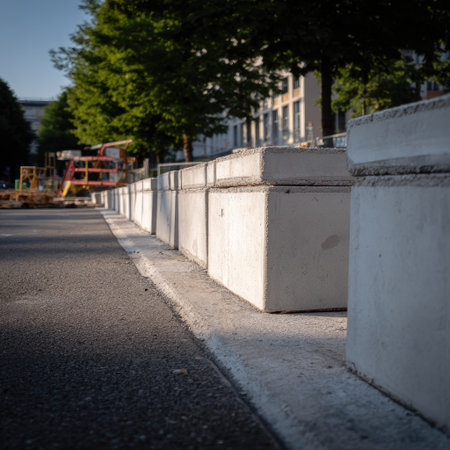 Concrete blocks form a barrier along an asphalt road, their gray surfaces contrasting with the dark road. The scene is bathed in natural light, suggesting an outdoor setting. The composition includes construction equipment in the background. This image is suitable for various commercial uses, including illustrating infrastructure and safety concepts.の素材