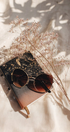 Close-up view of a notebook, round sunglasses, and a bouquet of delicate dried flowers on a light beige surface, bathed in the soft, warm light of the sun. The notebook features a subtle ombre pattern of brown and dark tones, while the sunglasses are a warm brown color. The dried flowers are a pastel pink hue.の素材