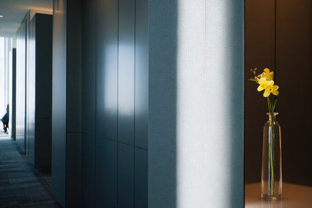 Single stem of yellow flowers in a clear glass vase stands in a modern hallway bathed in sunlight, highlighting the interplay of light and shadow on the smooth surfaces of the corridor.の素材
