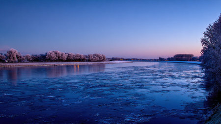 Wide, horizontal view of a large body of water, likely a river, at either dawn or dusk. The water surface is partially covered with sheets or chunks of ice, creating textures. The sky exhibits deep blue transitioning to soft pink and purple near the horizon. Trees lining the banks are heavily coated in white frost, contrasting with the dark water and sky. A few faint lights are visible on the far bank, reflecting slightly on the water.の素材