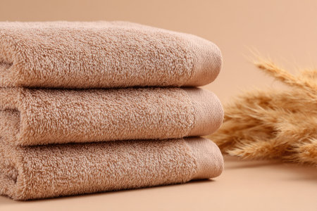 Stack of three beige terry cloth bath towels, arranged neatly on a soft, beige background.  A few dried grass stalks are visible in the background, adding to the overall neutral color scheme.の素材