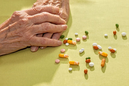 Clasped hands of an elderly lady resting on a table alongside a variety of scattered medication, tablets capsule and pills, prescribed for her health and as diet supplementsの写真素材