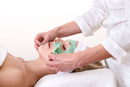 Beautician peeling off the green beauty facial mask on a laying and relaxed beautiful blond woman against a white backgroundの写真素材