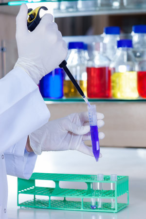 Hands of a lab technician using a pipette to transfer a measured amount of a blue solution from a...