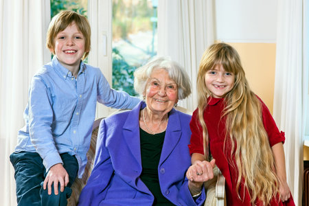 Smiling elderly grey-haired grandmother posing with her two grandchildren sitting flanked by a happy...