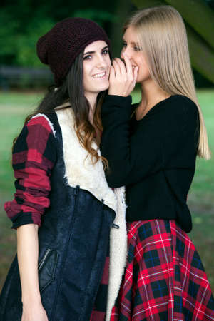 Two young female friends standing in a park talking and whispering, sharing secrets.の写真素材