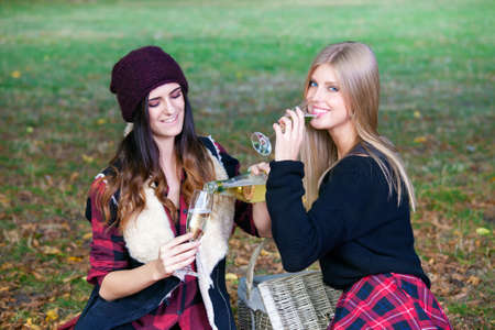Two beautiful girls having picnic in a park among autumn leaves, pouring champagne.の写真素材