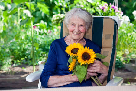 Close up Happy Old Lady in Blue Dress, Sitting on a Chair at the Garden, Holding Fresh Sunflowers While Looking at the Camera.の写真素材