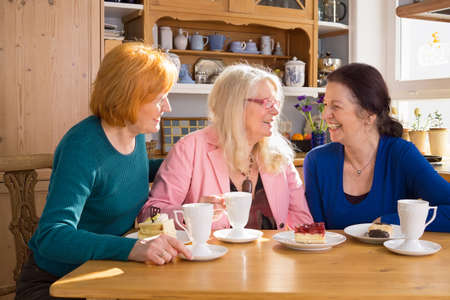 Three Happy Adult Female Friends Having Cups of Tea or Coffee and Slices of Cakes at the Wooden Table While Talking Funny Moments.の写真素材