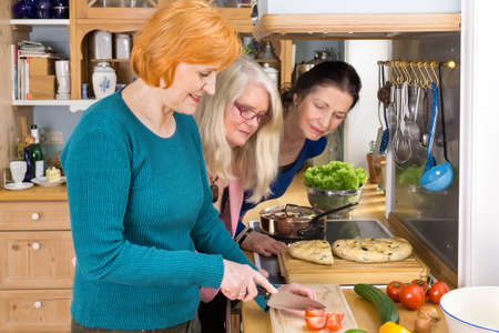 Curious Middle Age Moms Looking at their Smiling Friend Slicing Ingredients for their Dinner Recipe.の写真素材