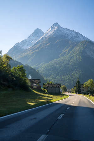 Quaint Houses Along Paved Mountain Road Through Lush Green Valley with Snow Capped Alps Valais Switzerland on Sunny Day.の写真素材