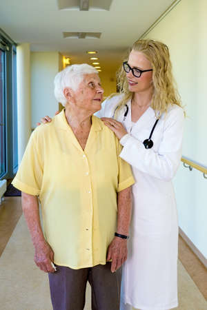 Three-Quarter Shot of a Kind Young Nurse Talking to an Elderly Woman with Special Needs at the Corridor Inside the Nursing Home.の写真素材