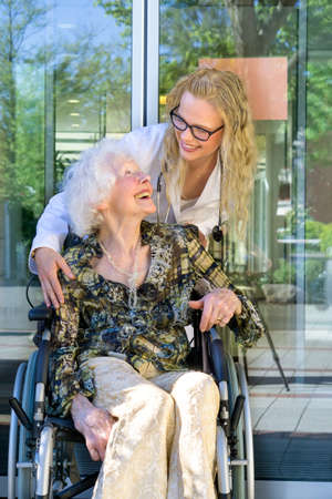 Young Medical Assistant and her Elderly Woman Patient on a Wheelchair Laughing Together Outside the Nursing Home.の写真素材
