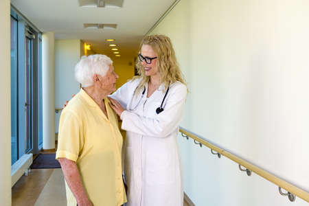 Cheerful Young Nurse Assisting a Senior Woman with Special Needs Walking at the Corridor Inside the Hospital.の写真素材