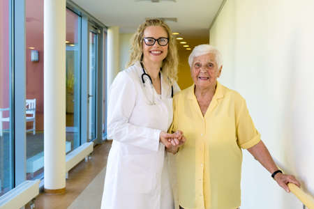 Kind Female Nurse Assisting her Old Woman Patient at the Hallway in the Hospital, Smiling at the Cameraの写真素材