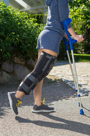 Young woman wearing an adjustable leg brace to support and immobilize her knee post operative, side view, walking outdoors on crutches in a gardenの写真素材