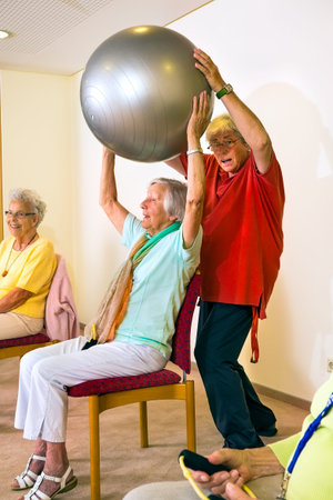 Fitness trainer working with smiling senior woman lifting a silver stability ball for exercise at an old-age homeの写真素材