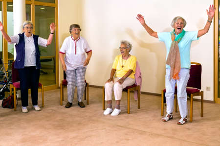 Group of four cheering senior women practicing light aerobic exercises with chairs for fitness class indoorsの写真素材