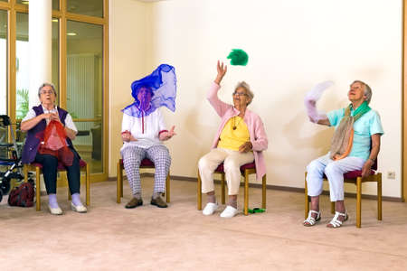 Senior ladies having fun while exercising at a seniors gym as they sit on chairs throwing colorful scarves in the air to improve coordinationの写真素材