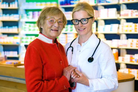 Happy older woman holding hands with smiling young female doctor in eyeglasses and stethoscope standing in front of pharmacy counterの写真素材