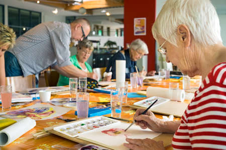 Woman in striped red and white shirt working on canvas while painting with brush at table with other students and teacher in spacious studioの写真素材