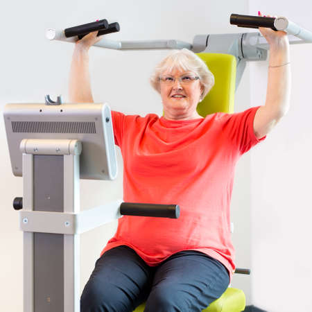 Smiling single senior woman using shoulder machine to improve upper body strength in fitness areaの写真素材