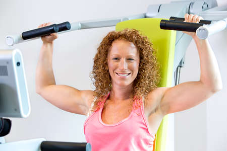 Cheerful athletic female in red curly hair and pink top with arms in the lowered position with shoulder strengthening machineの写真素材
