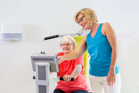 Friendly female trainer in eyeglasses and blue shirt helping senior patient with settings on exercise machineの写真素材