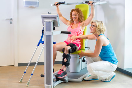 Patient in exercise machine and orthopedic brace getting help with leg rehabilitation from...