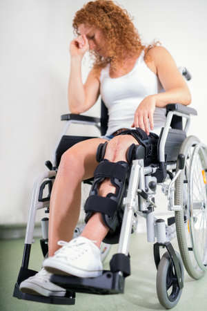 Dejected woman with her knee in a brace and a post-operative scar sitting in a wheelchair with her head resting on her hand in a disability and healthcare conceptの写真素材