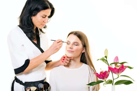 Beautician applying eye makeup to a pretty young woman in a spa or beauty salon using a small cosmetics brush with fresh pink lilies in the foreground isolated on white with copy spaceの写真素材