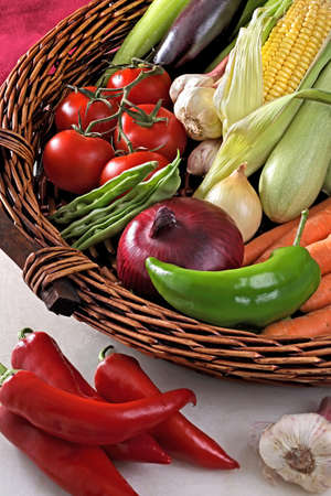 Brown wicker basket with variety of fresh vegetables viewed in close-up from high angle  with red peppers and garlic on white table.の写真素材
