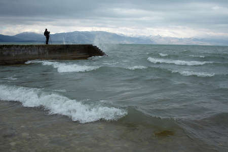 Woman standing on concrete pier at wavy sea under cloudy sky, viewed from the side. Waves breaking on the shore in foreground.の写真素材