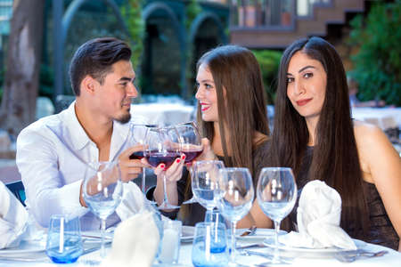 Three friends having dinner at an open air restaurant with two elegant young women and a man toasting each other with glasses of red wine at a formal table.の写真素材