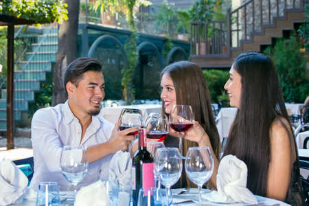 Handsome man with two beautiful women in restaurant sitting at table and clinking glasses of red wine, looking at each other and smiling.の写真素材